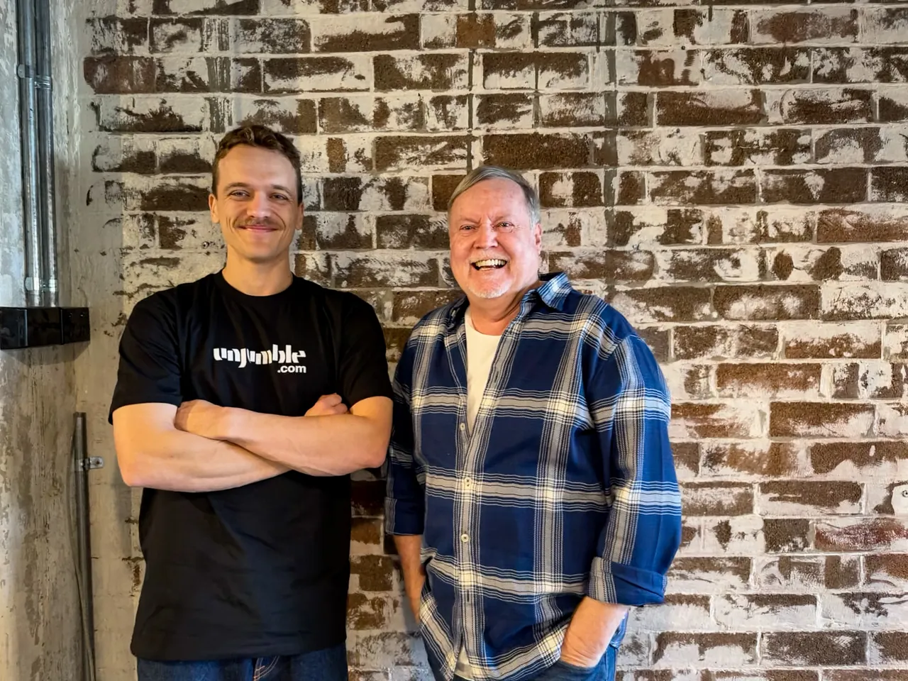 Zac Kelly in an Unjumble t-shirt standing beside Greg "Sparrow" Graham in front of a whitewashed brick wall